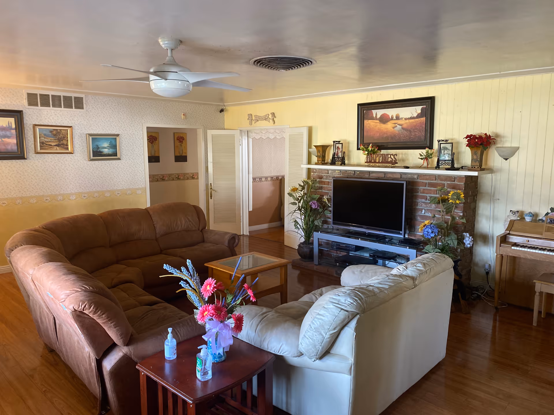 A cozy living room with a brown sectional sofa and a white armchair arranged around a glass coffee table. There is a wooden side table with a vase of colorful flowers and hand sanitizer bottles. The room features a ceiling fan, framed paintings on the walls, a brick fireplace with a TV on a stand, decorative plants, and a piano in the corner.