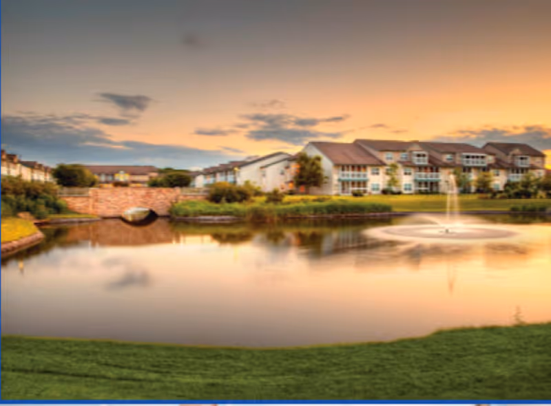 A serene view of a senior living facility at sunset, featuring a large pond with a water fountain in the center, surrounded by well-maintained green lawns and residential buildings in the background under a partly cloudy sky.