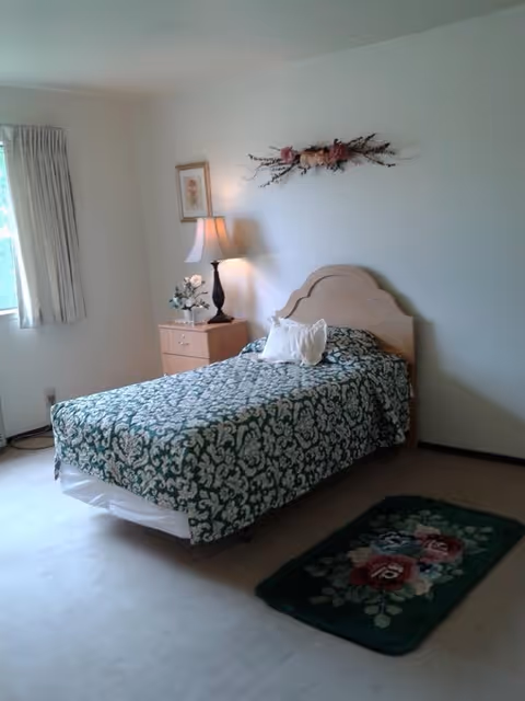 A simple bedroom with a single bed covered in a green and white patterned bedspread. There is a wooden headboard, a white pillow, and a small wooden nightstand with a lamp and a flower arrangement. A framed picture hangs on the wall above the nightstand, and a floral wall decoration is above the headboard. A window with light-colored curtains is on the left side, and a small floral rug is on the carpeted floor near the foot of the bed.