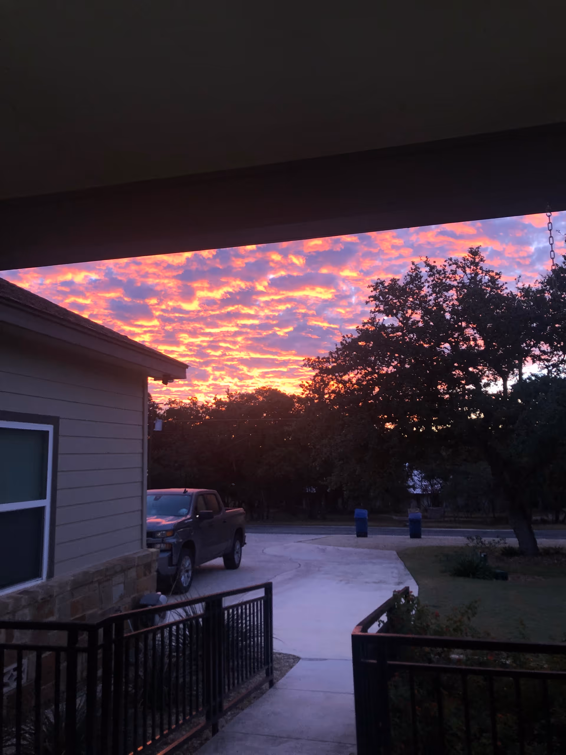 View from a covered porch or patio showing a driveway with a parked pickup truck, a house on the left, trees in the background, and a vibrant sunset with pink and orange clouds in the sky.