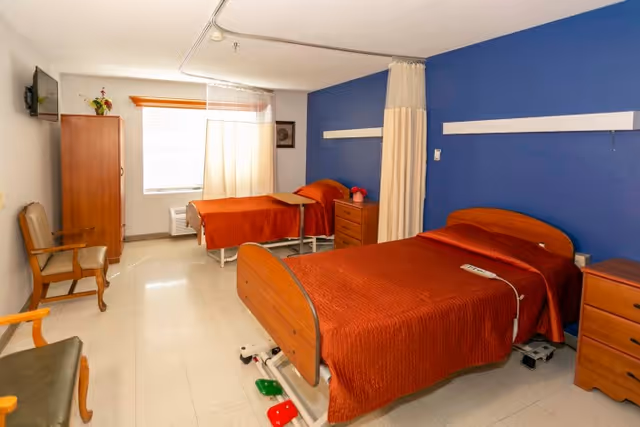 A bright room with two hospital-style beds covered in orange bedspreads. Each bed has a wooden headboard and footboard, and there are wooden nightstands beside each bed. A curtain divider hangs from the ceiling between the beds. The room has a blue accent wall behind the beds and a window with white curtains letting in natural light. There are two wooden chairs and a wooden cabinet along the opposite wall, and a flat-screen TV mounted on the wall.