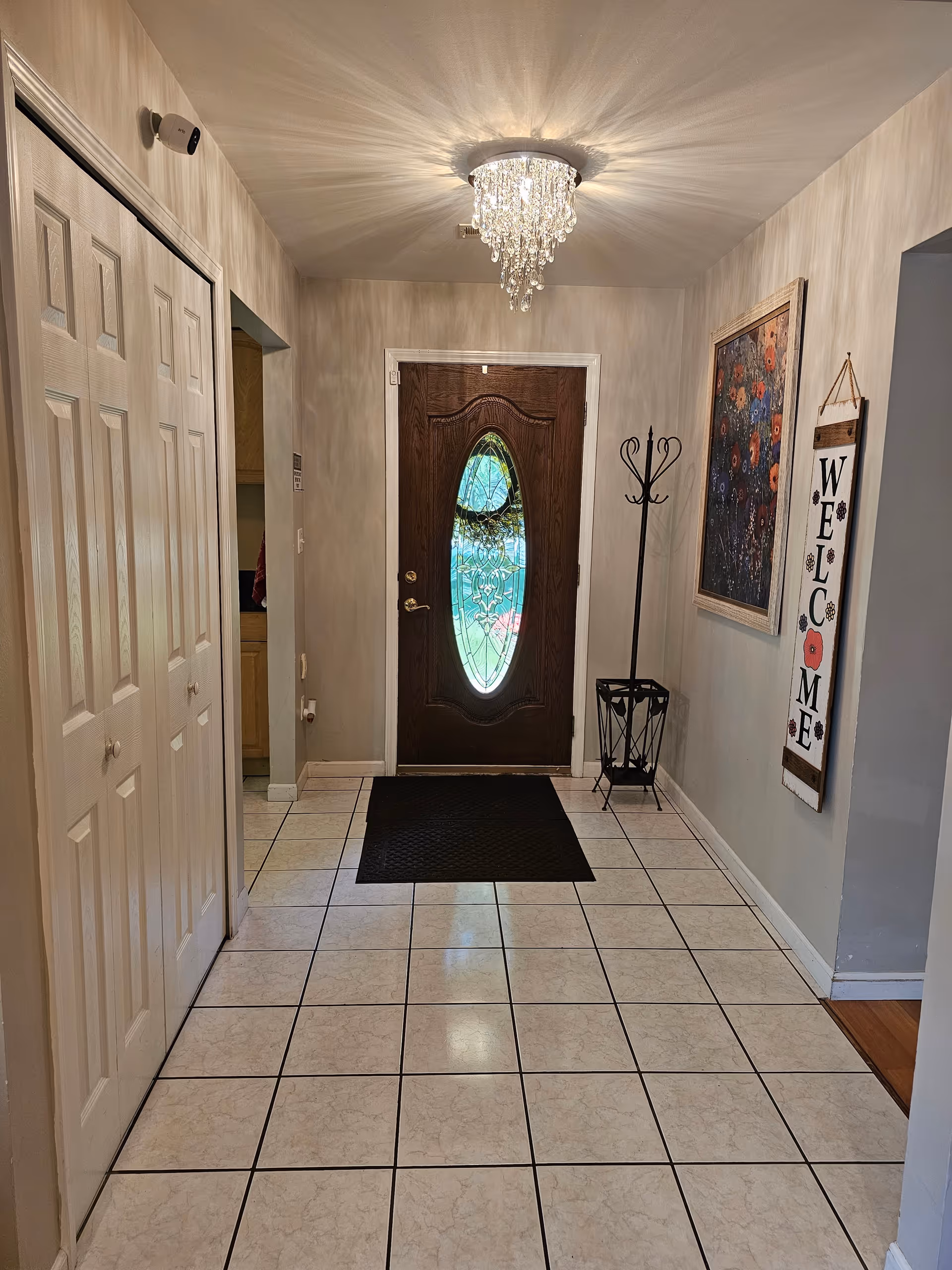Entryway of a senior care home with a decorative wooden front door featuring an oval glass panel. The floor is tiled with white tiles and black grout. A black mat is placed in front of the door. On the right wall, there is a colorful floral painting and a vertical wooden welcome sign. A black coat rack and umbrella stand are also visible. The ceiling has a crystal chandelier light fixture.