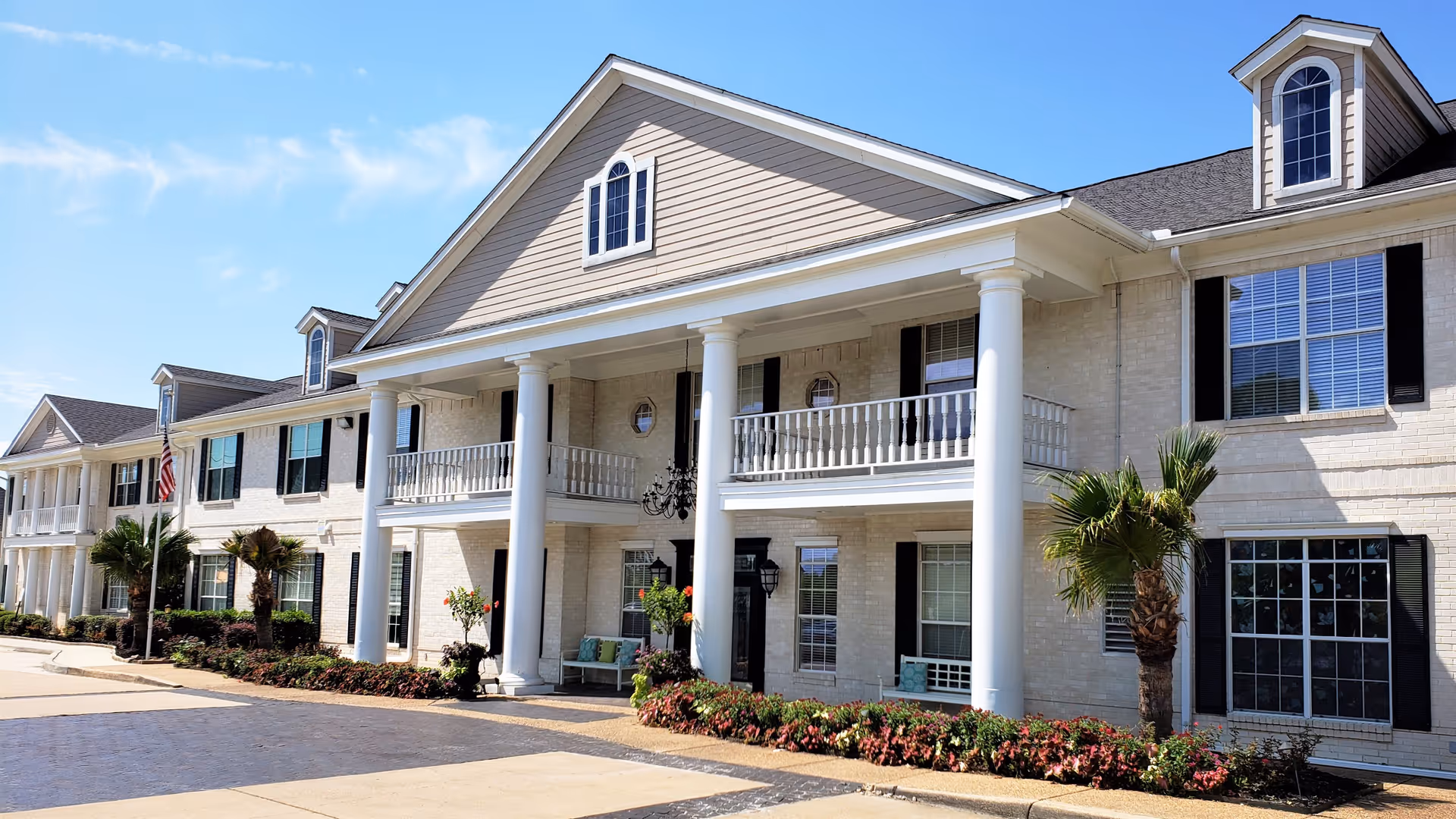 Front exterior view of Montclair Park Assisted Living building with white columns, balconies, large windows with black shutters, palm trees, and landscaped flower beds under a clear blue sky.