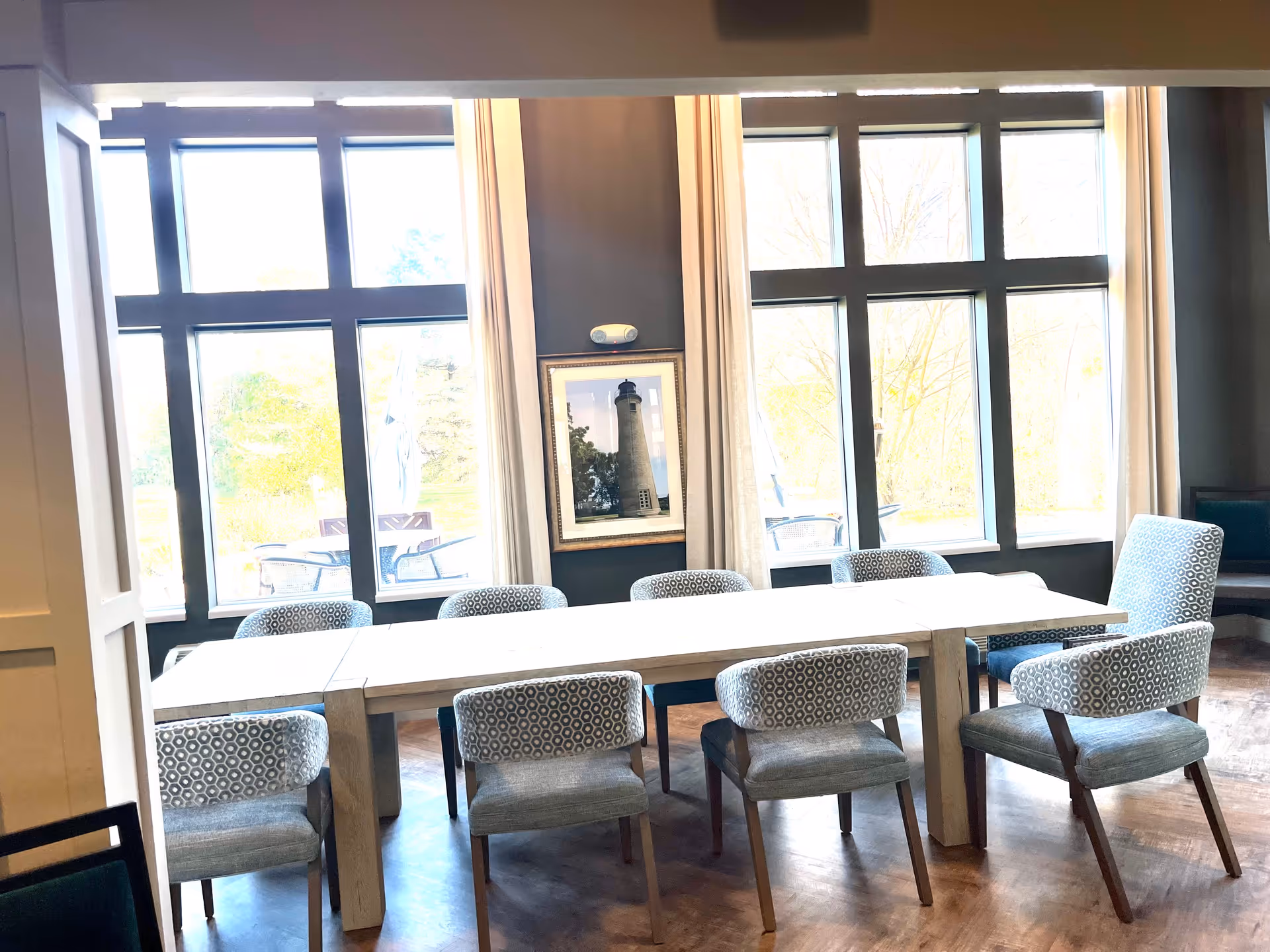 A bright dining room with a long rectangular table surrounded by eight patterned chairs. Large windows with white curtains let in natural light, and a framed picture of a lighthouse hangs on the dark wall between the windows. Outside, patio furniture is visible through the windows.