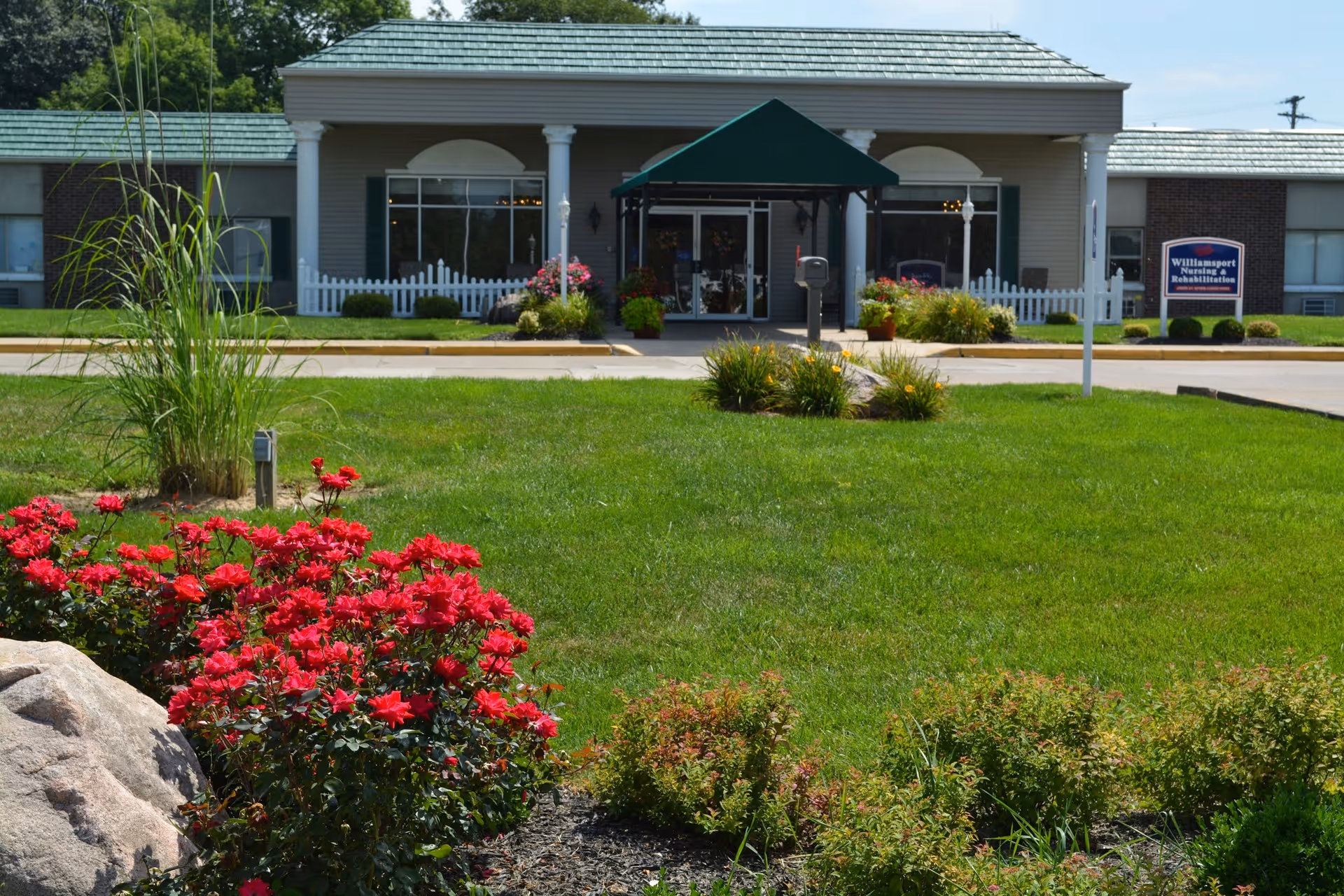 Front entrance of Williamsport Nursing and Rehabilitation with white columns, a green canopy, and a manicured lawn with red flowers in the foreground.