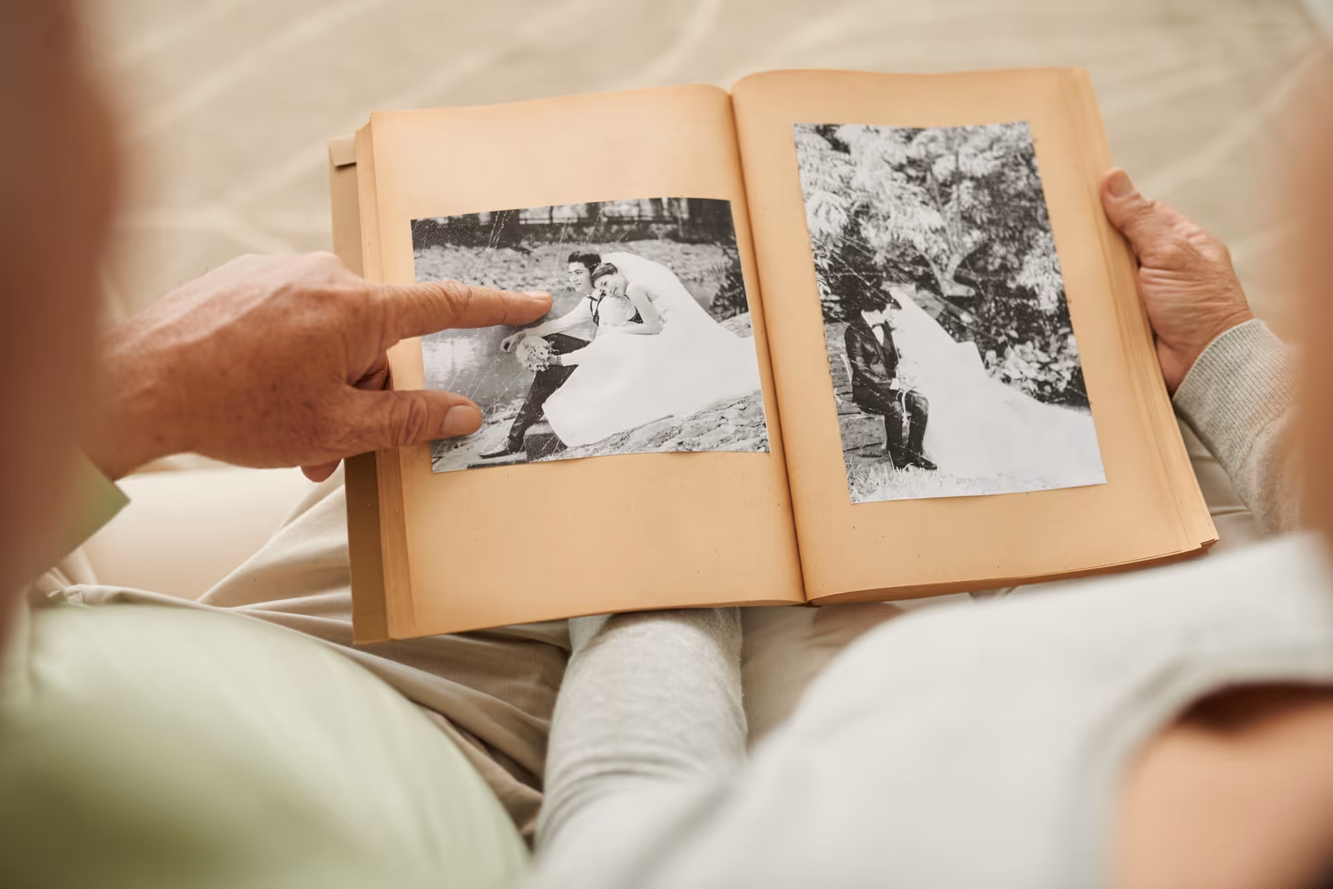 Two elderly people sitting closely together looking at a photo album with black and white wedding photos. One person is pointing at a photo of a bride and groom sitting by a pond.