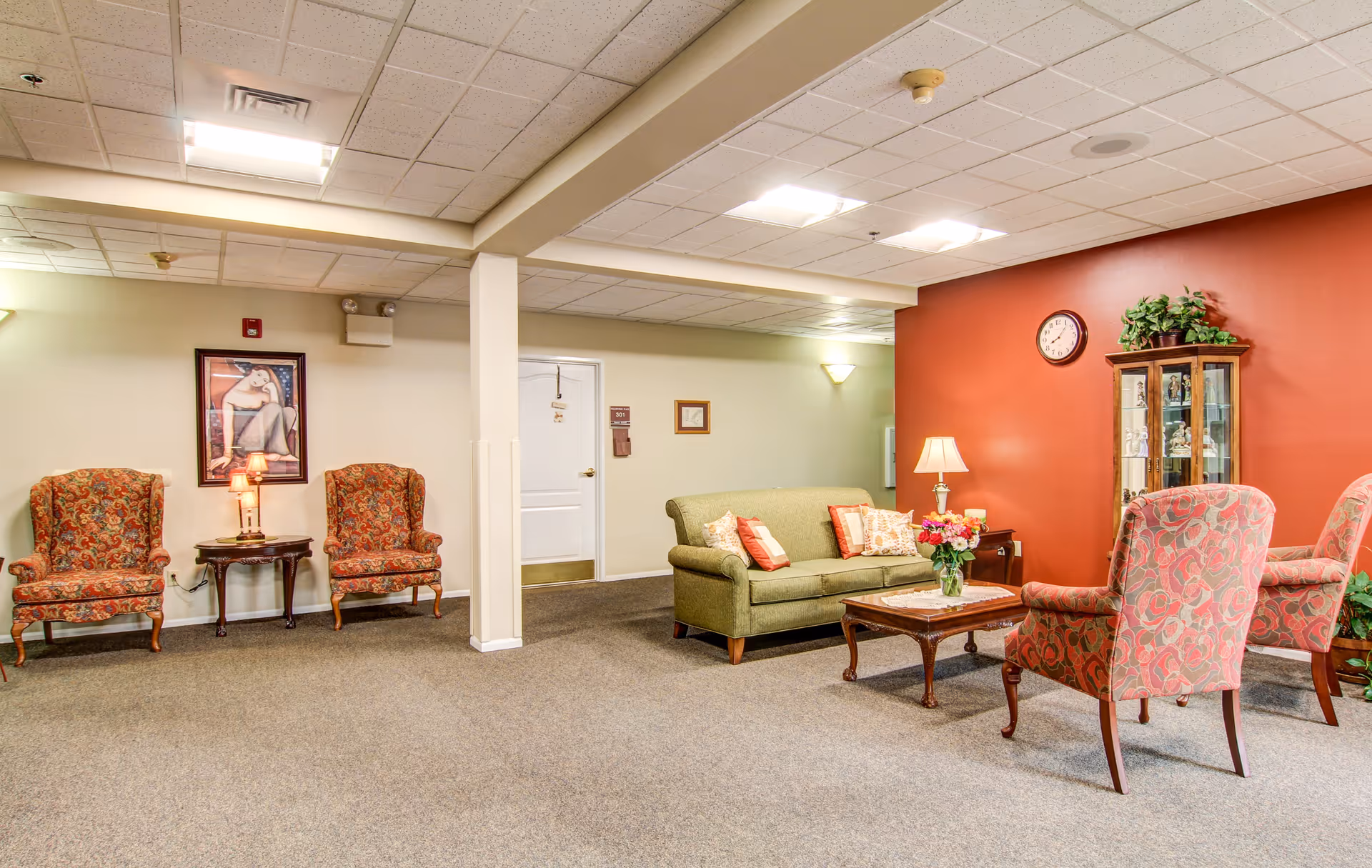 A cozy living room area in Richmond Terrace featuring a green sofa with decorative pillows, a wooden coffee table with a flower vase, two patterned armchairs, and a wooden display cabinet against a red accent wall. There are also two patterned chairs with a small table and lamp between them on the opposite side of the room. The ceiling has recessed lighting and the floor is carpeted.