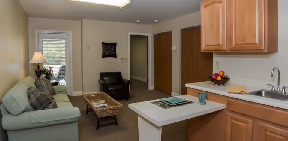 A cozy living area in a senior living facility featuring a light green sofa with plaid pillows, a wicker coffee table with magazines, a black armchair, and a small kitchen area with wooden cabinets, a sink, and a countertop with a bowl of fruit and a cup. A door with blinds leads to an outdoor space.