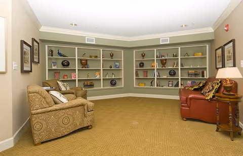 A cozy living room area with patterned armchairs and a red leather armchair arranged around a corner with built-in shelves displaying decorative items and vases. The room has beige walls, carpeted floor, framed pictures on the walls, and a table lamp on a small wooden side table.