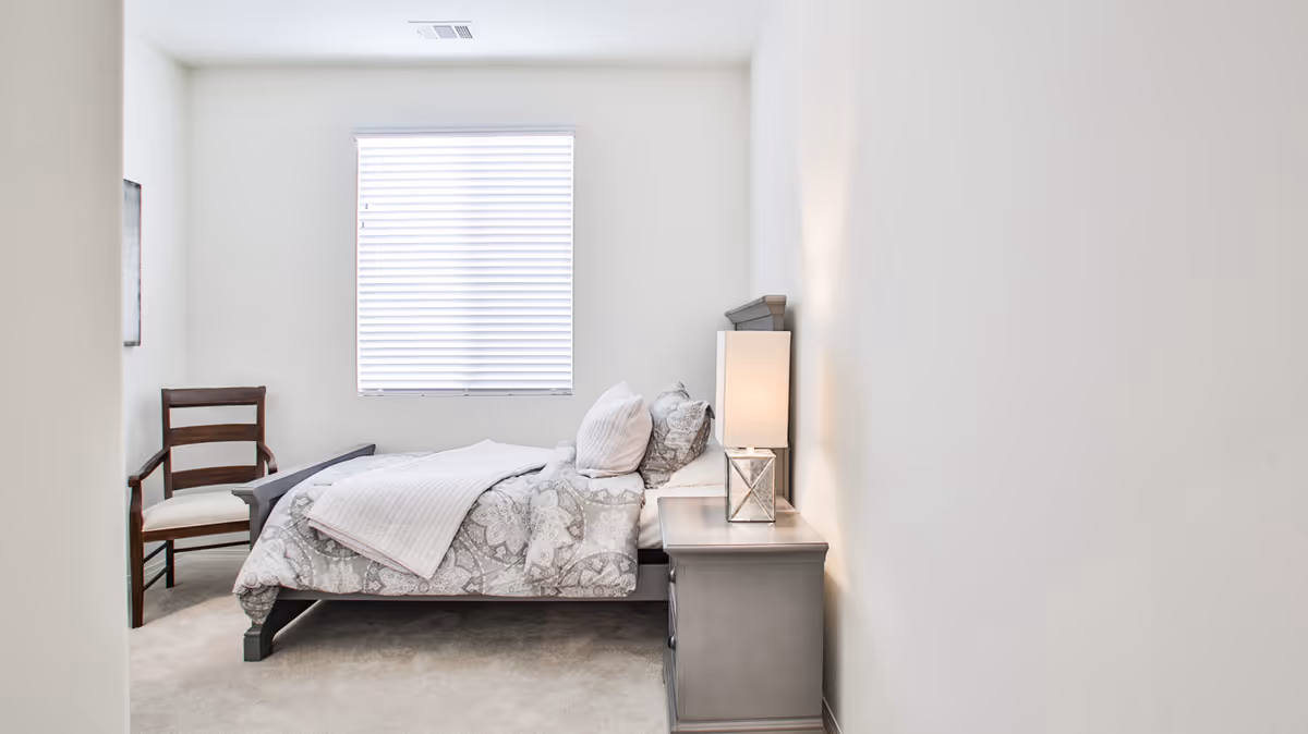 A simple, bright bedroom with a bed dressed in patterned bedding, a nightstand with a lamp, a wooden chair, and a window with blinds.