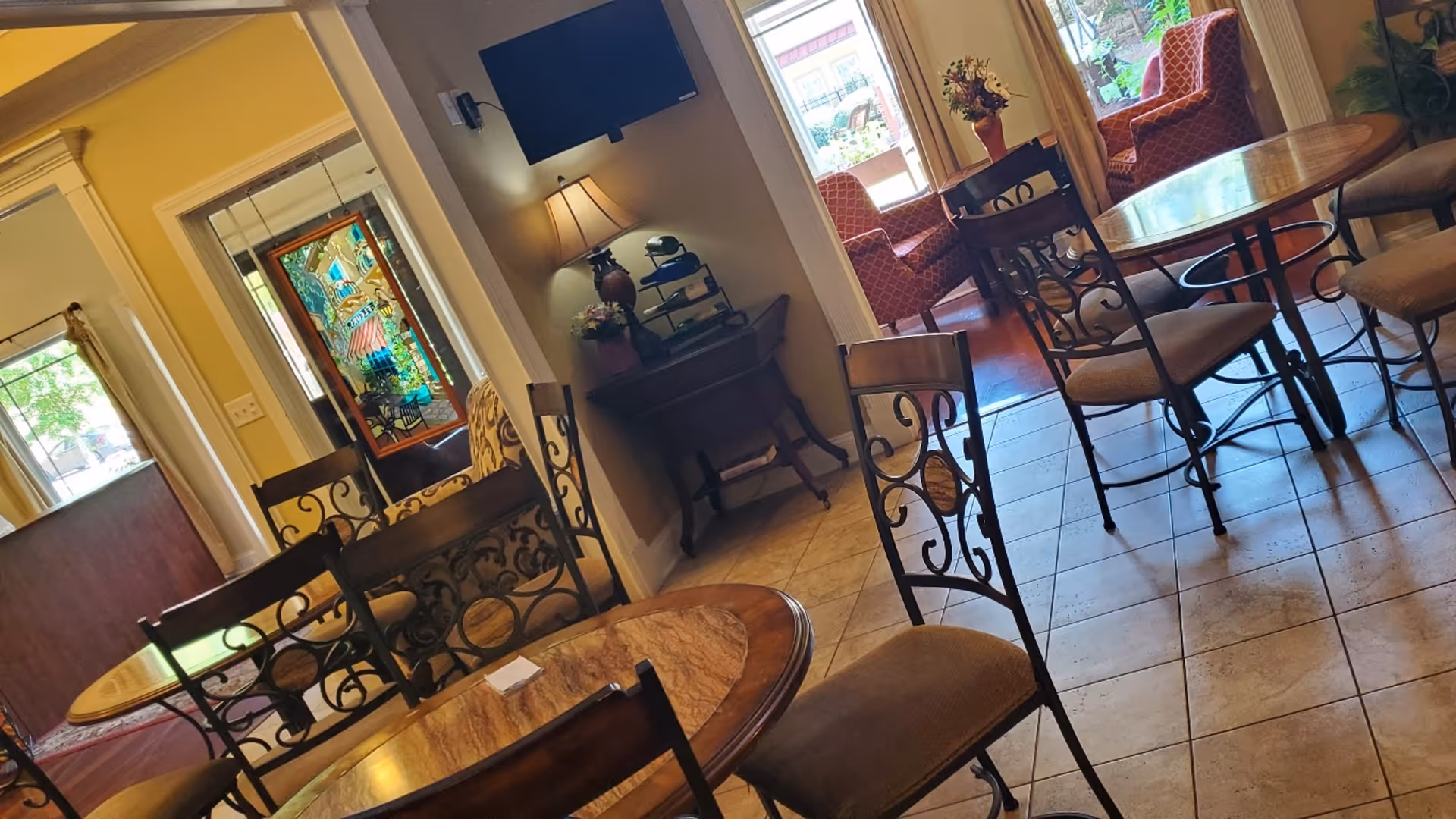 Cozy assisted-living common area with round tables, wrought-iron chairs, a lamp on a side table, and upholstered seating by windows.