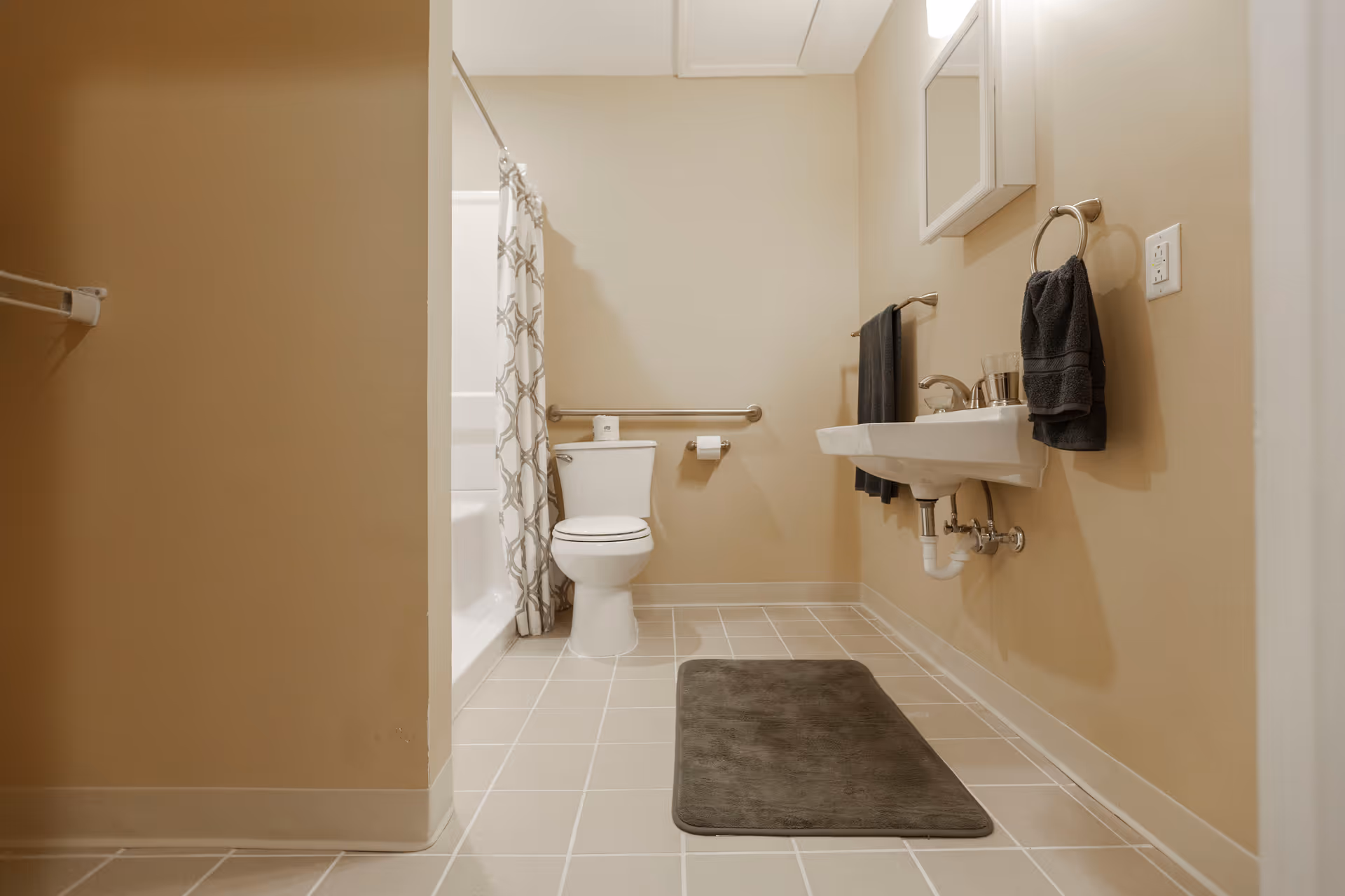 A clean bathroom with beige walls and tiled floor. The room features a white toilet with a grab bar next to it, a bathtub with a patterned shower curtain, a wall-mounted sink with a faucet, a mirror above the sink, and two dark gray towels hanging on towel racks. A dark gray bath mat is placed on the floor in front of the sink.