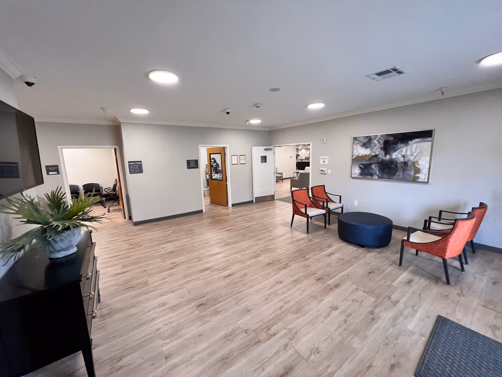 A spacious waiting or common area with light wood flooring, four red cushioned chairs arranged around a round black ottoman, a large abstract painting on the wall, and a black cabinet with a potted plant. There are multiple doorways leading to other rooms, and the ceiling has several round recessed lights.