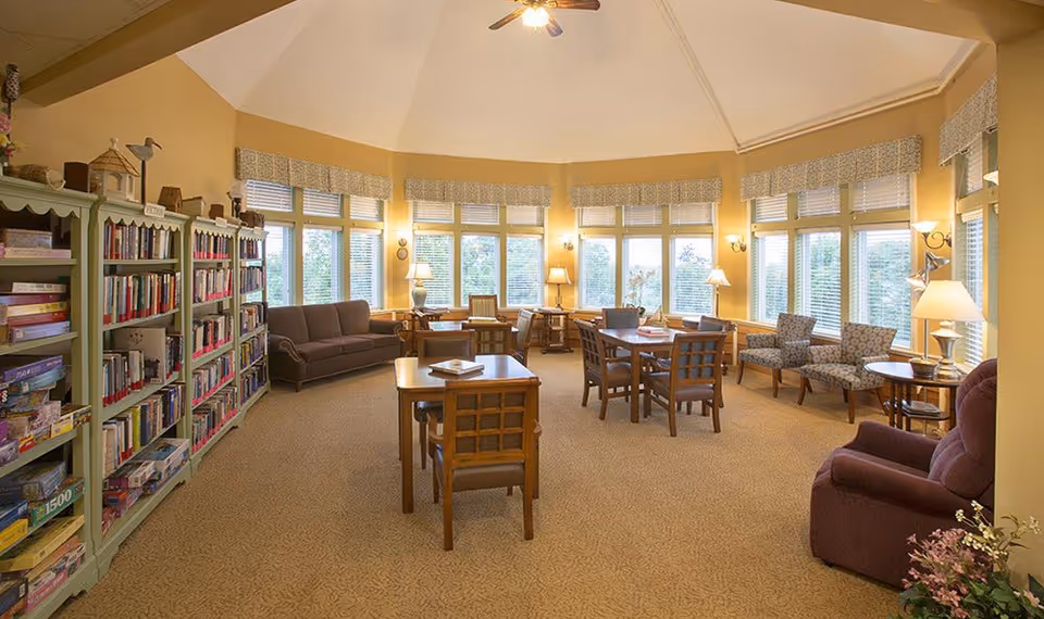 A spacious and well-lit common area with large windows letting in natural light. The room features multiple wooden tables and chairs, a bookshelf filled with books and board games, several armchairs and sofas, and various lamps providing warm lighting. The walls are painted a soft yellow, and the carpet is beige with a subtle pattern.
