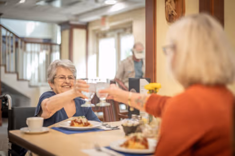 Two elderly women sitting at a dining table in a senior living facility, smiling and clinking their glasses in a toast. Plates of food and a cup are on the table, with a bright and welcoming interior in the background.