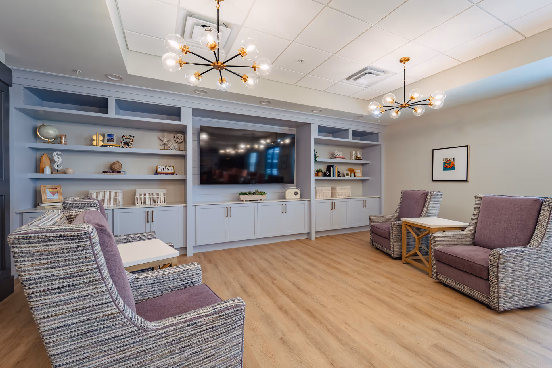 A modern living room with four upholstered armchairs featuring purple cushions arranged around two small wooden tables. The room has light wood flooring, built-in light blue shelves and cabinets along one wall, and a large flat-screen TV mounted in the center. Decorative items and books are displayed on the shelves. Two stylish chandeliers with multiple globe lights hang from the ceiling, and a framed artwork is on the wall.