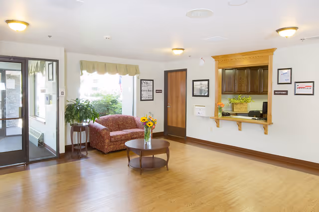 A bright reception area with a wooden floor, a floral-patterned loveseat, a round wooden coffee table with a vase of flowers, a plant on a small table near a large window, and a reception window with a wooden frame on the right wall. There is a door next to the reception window and glass entrance doors on the left side.
