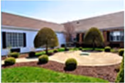Outdoor courtyard area with trimmed bushes, small trees, and a circular paved section surrounded by mulch and grass, adjacent to a single-story building with white walls and windows.