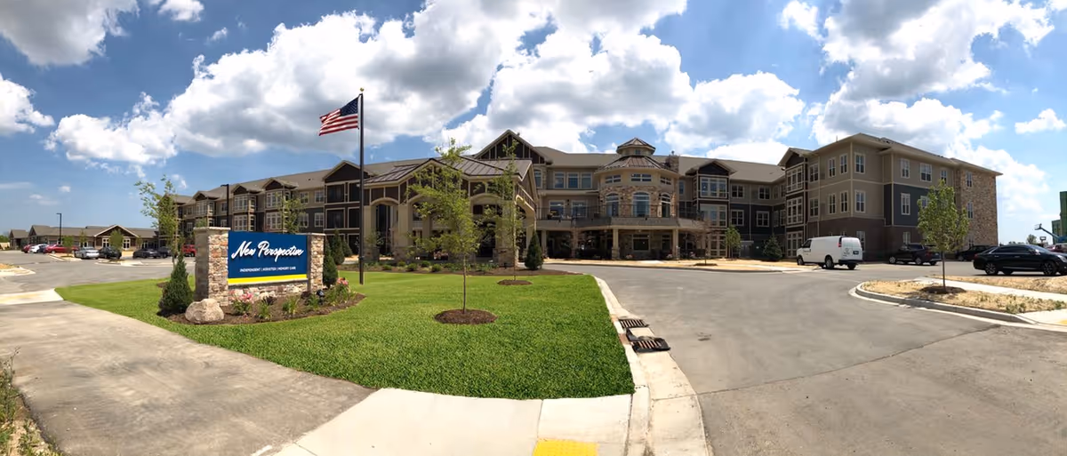Wide exterior view of New Perspective Senior Living | Franklin building under a partly cloudy sky, with a green lawn, an American flag on a pole, a sign displaying the facility name, and a parking area with several vehicles.