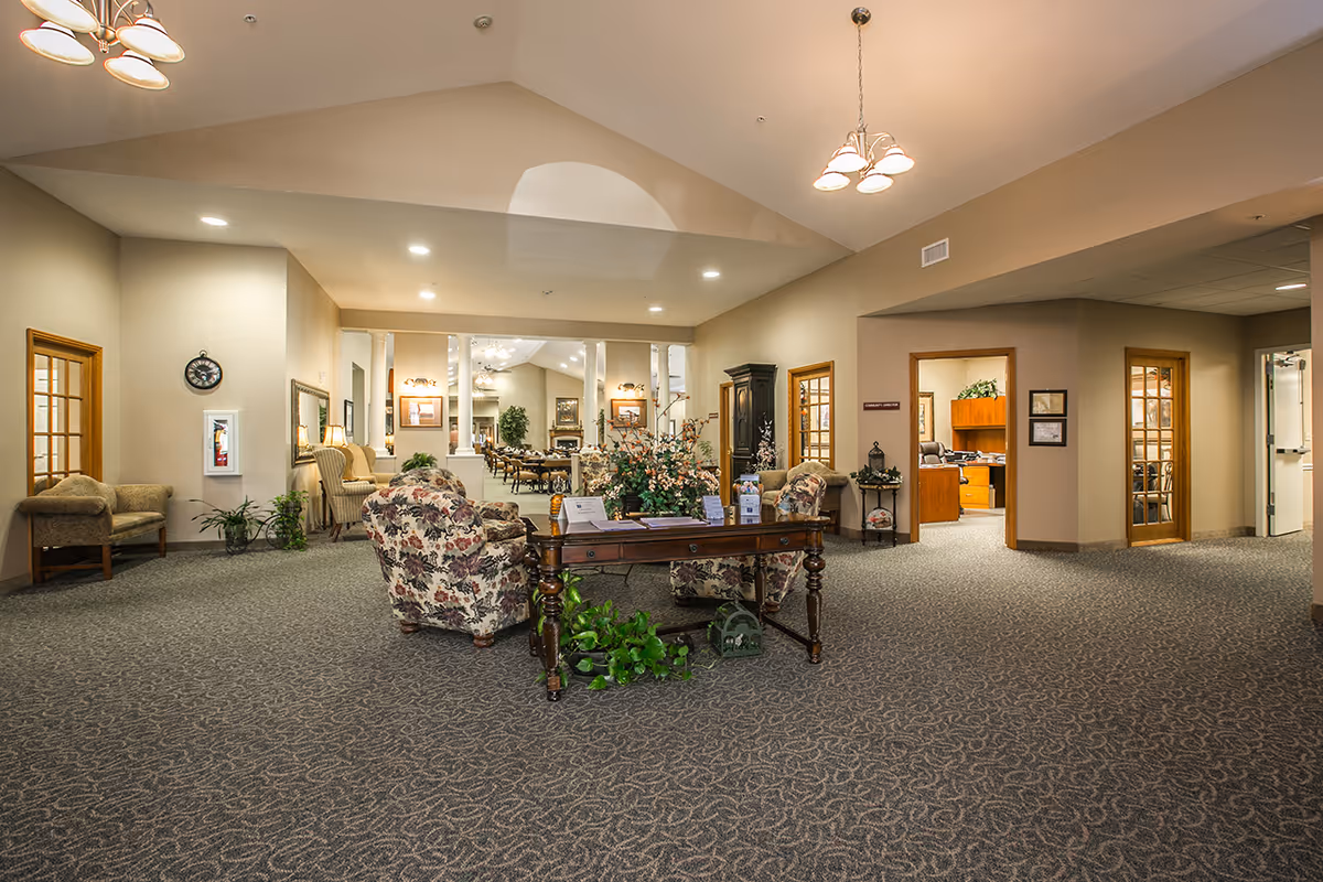 Spacious and well-lit interior common area of The Bungalows at Fayetteville featuring floral upholstered armchairs around a wooden table with a flower arrangement, carpeted floor, beige walls, multiple doorways leading to other rooms, and ceiling lights.