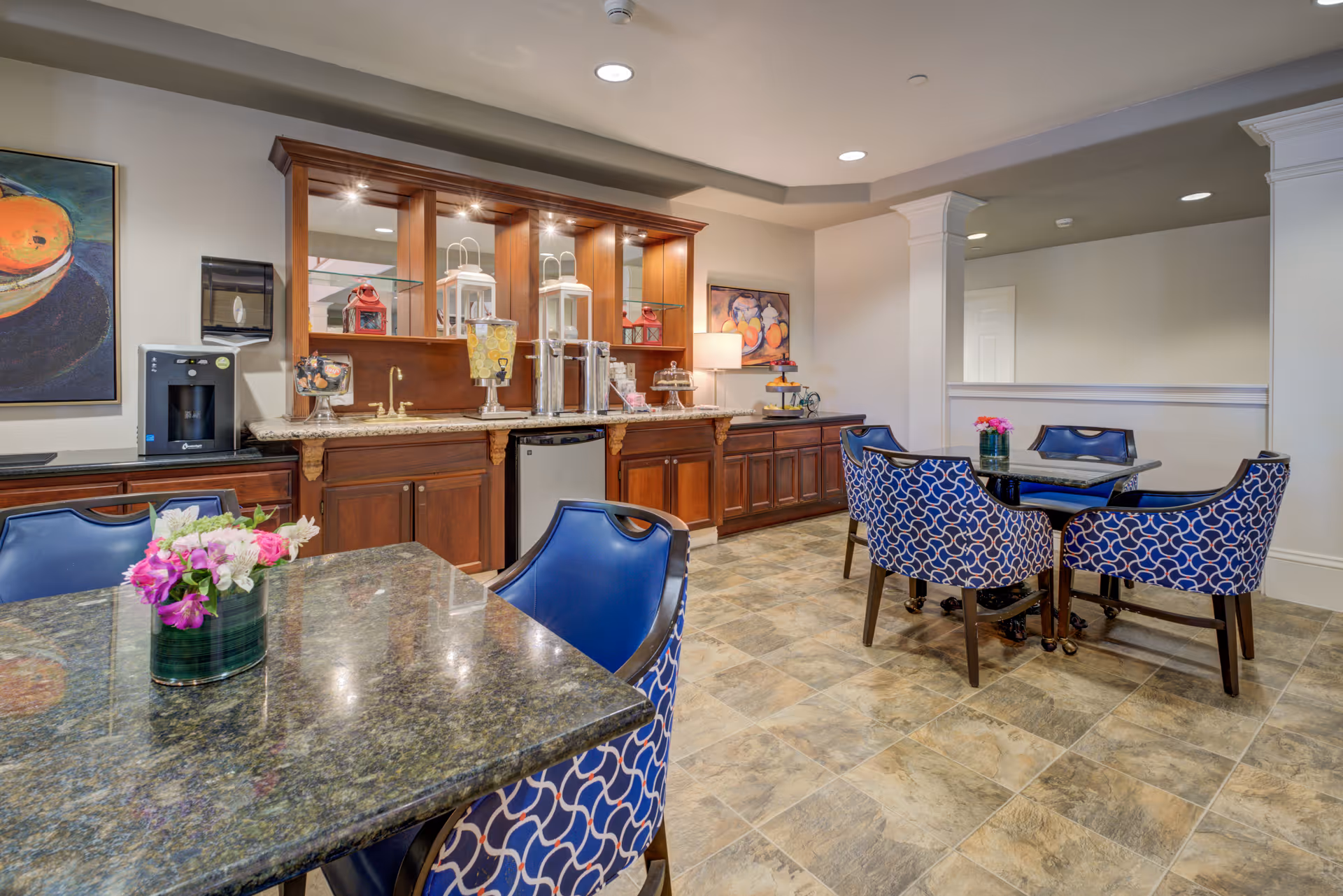 A cozy dining area in a senior living facility featuring two tables with blue patterned chairs. The tables have granite tops and small flower arrangements. In the background, there is a wooden cabinet with a countertop holding beverage dispensers, a coffee machine, and decorative lanterns. The room has tiled flooring and soft lighting, with paintings on the walls.