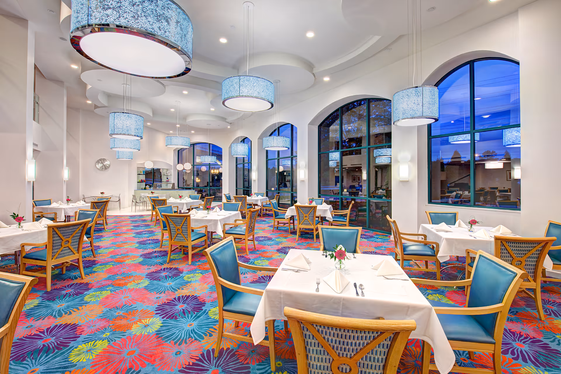 Bright and spacious dining room with large arched windows, colorful floral carpet, multiple tables covered with white tablecloths, each set with napkins, cutlery, and small flower vases. Blue cushioned wooden chairs surround the tables, and modern circular pendant lights hang from the ceiling.