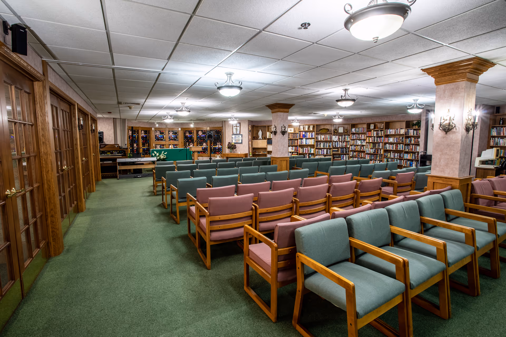 Interior view of a senior community room with rows of wooden chairs upholstered in green and pink fabric facing a small stage area with a green podium. The room has green carpet, wooden columns, bookshelves filled with books along the walls, and ceiling lights.