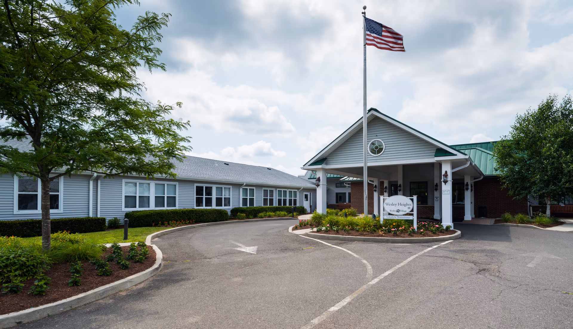 Exterior view of Masonicare at Wesley Heights facility showing a curved driveway leading to a covered entrance with a flagpole flying an American flag. The building has gray siding, white trim, and a green roof with landscaped bushes and trees around.