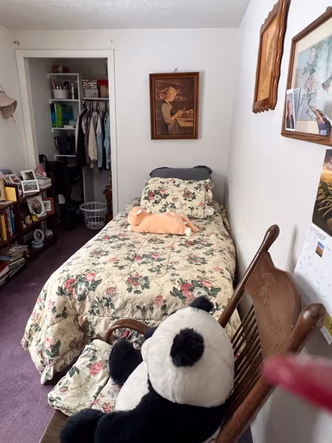A small bedroom with a single bed covered in a floral bedspread. A stuffed animal is placed on the bed. There is a wooden chair with a large stuffed panda bear sitting on it. The room has a purple carpet and white walls decorated with framed pictures. An open closet with hanging clothes and shelves is visible at the far end of the room.