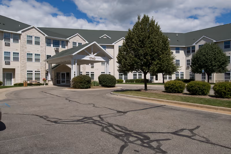 Exterior view of a senior living facility building with a covered entrance marked 1740, surrounded by trees and bushes under a partly cloudy sky.