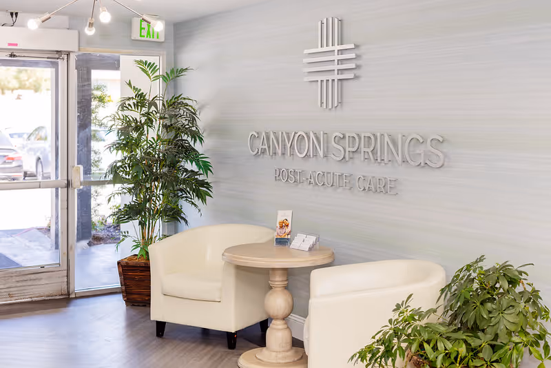 Reception area of Canyon Springs Post-Acute Care featuring two white armchairs around a small round wooden table with brochures, potted plants, and a glass door entrance with an exit sign above.