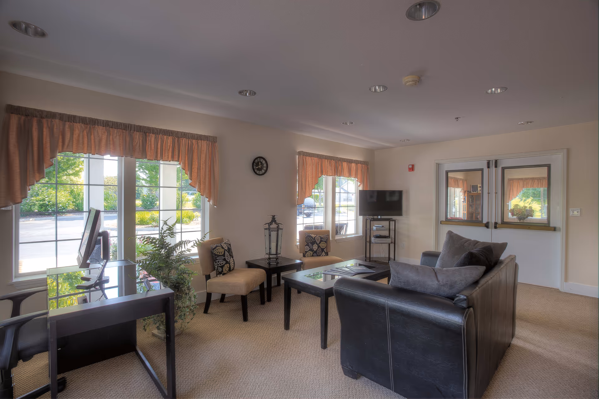 A cozy living room area in a senior living facility with a black leather couch, two beige chairs with patterned cushions, a glass coffee table, a small TV on a stand, a desk with a computer, and large windows with peach-colored valances letting in natural light.
