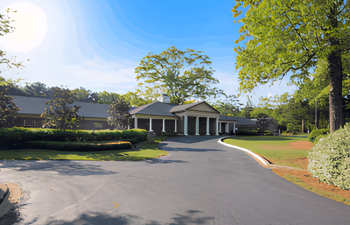 Exterior view of a single-story building with a covered entrance supported by columns, surrounded by green lawns, bushes, and trees under a clear blue sky with the sun shining.