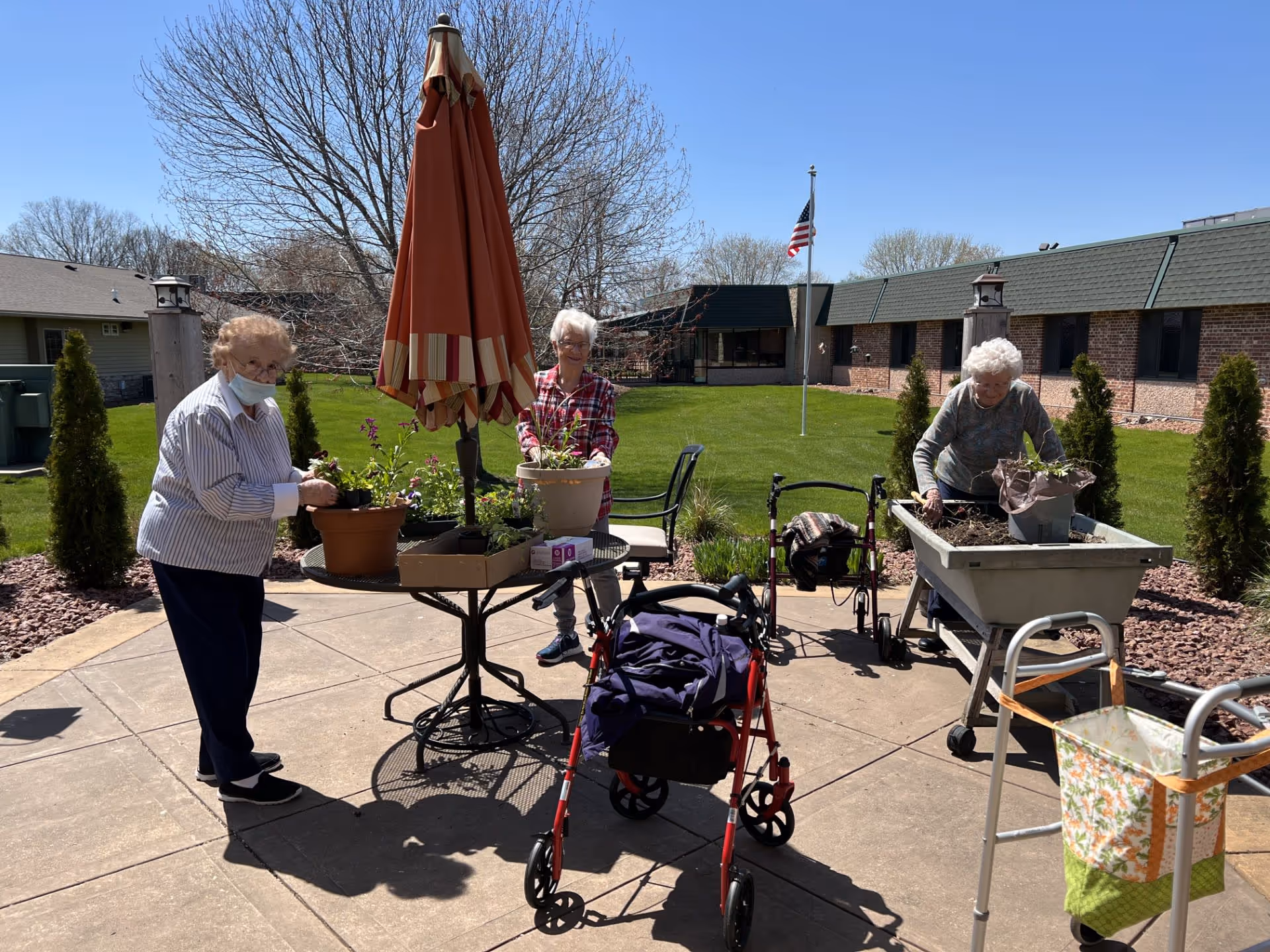 Three older adults tending potted plants around a patio table with an umbrella in a grassy courtyard, with walkers and a building in the background.