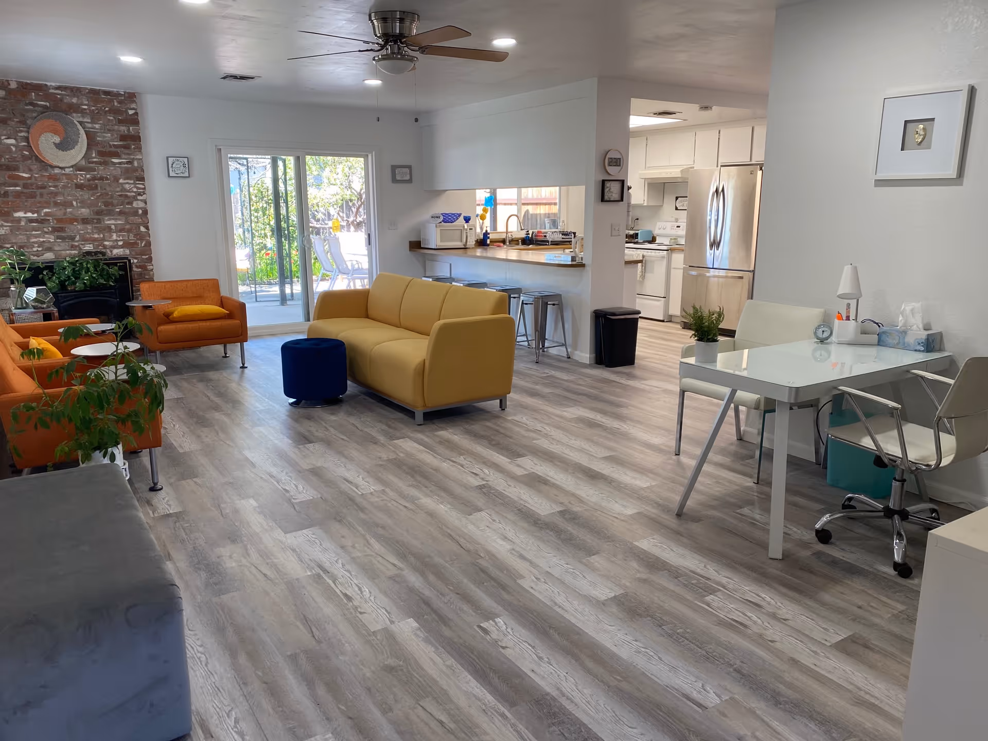 A bright and spacious living area with light wood flooring, featuring a yellow sofa, two orange armchairs, a blue ottoman, and a gray bench. There is a brick fireplace on the left wall, a ceiling fan above, and sliding glass doors leading to an outdoor patio. To the right, there is a white desk with a chair and a small plant, and beyond that, a kitchen area with white cabinets and stainless steel refrigerator.