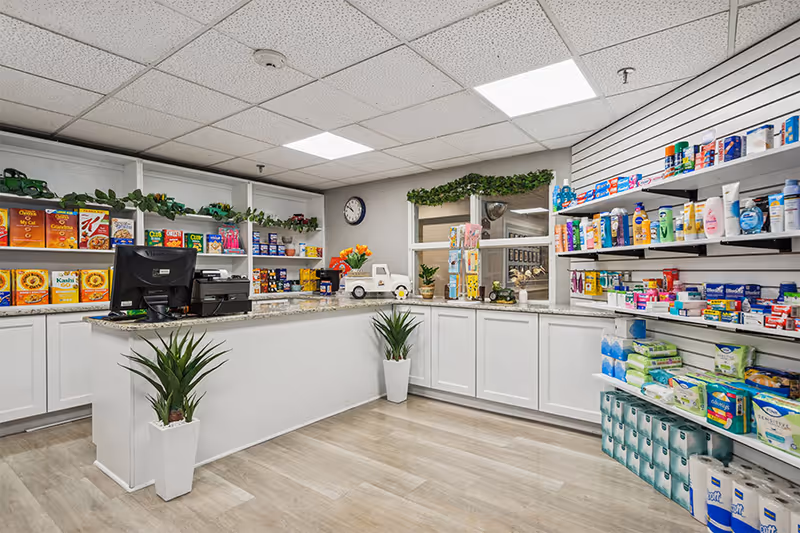 Interior view of a well-organized retail or convenience area in a senior living facility named Coral Oaks, featuring white cabinetry and shelves stocked with various grocery items, personal care products, and household essentials. The counter has a computer and a small decorative plant, with two potted plants on the floor. The ceiling has recessed lighting and a clock is mounted on the wall.