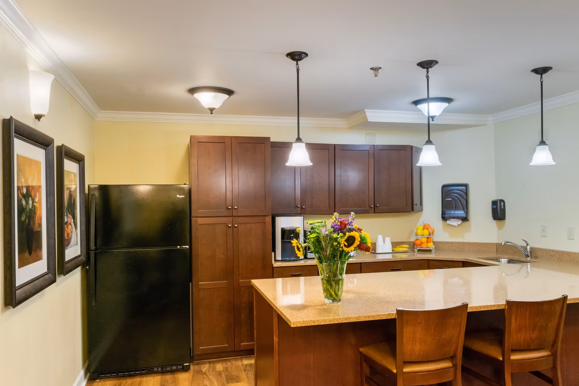 A kitchen area with wooden cabinets, a black refrigerator, a countertop island with two wooden chairs, and a vase of colorful flowers on the counter. The kitchen is well-lit with ceiling lights and pendant lamps. There are two framed pictures on the wall to the left.