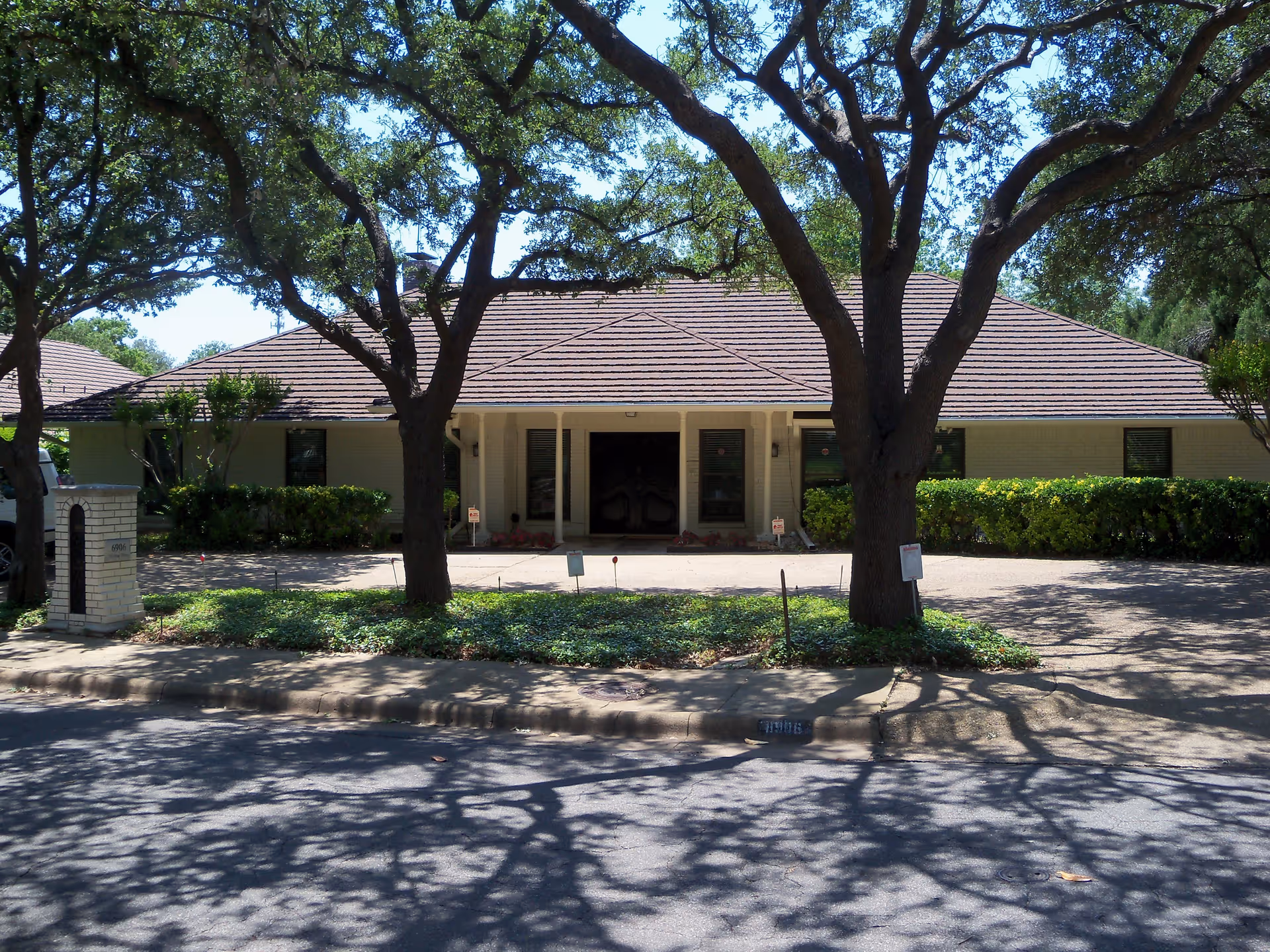 Front exterior view of a single-story building with a tiled roof, surrounded by trees and shrubs. The driveway and entrance are visible, with shadows of tree branches cast on the street in front.