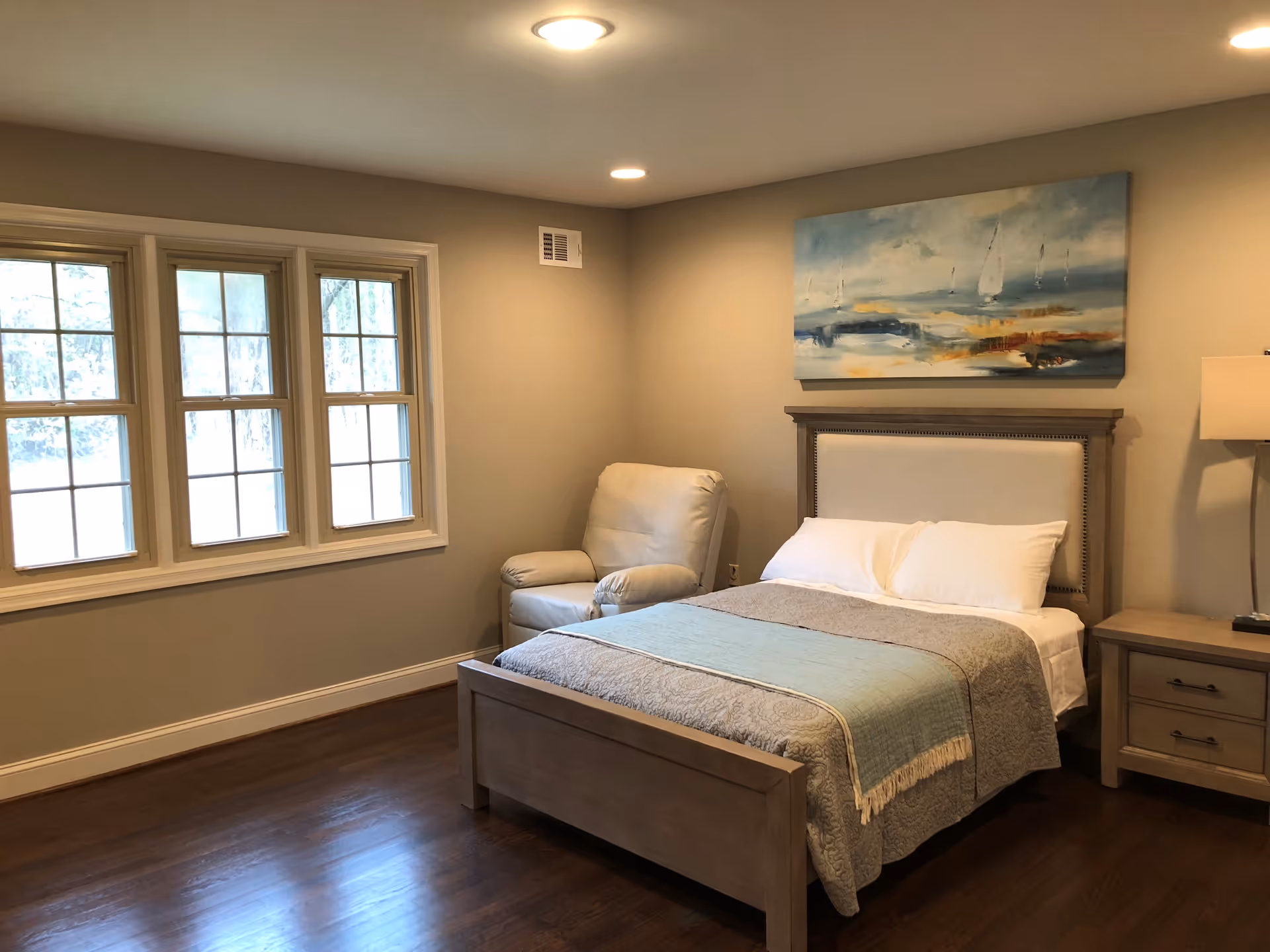 A bedroom with a wooden bed frame, a neatly made bed with white pillows and a gray and blue blanket. There is a beige armchair next to the bed, a wooden nightstand with a lamp, and a large abstract painting with blue and orange tones hanging on the wall above the bed. The room has wooden flooring and three large windows letting in natural light.