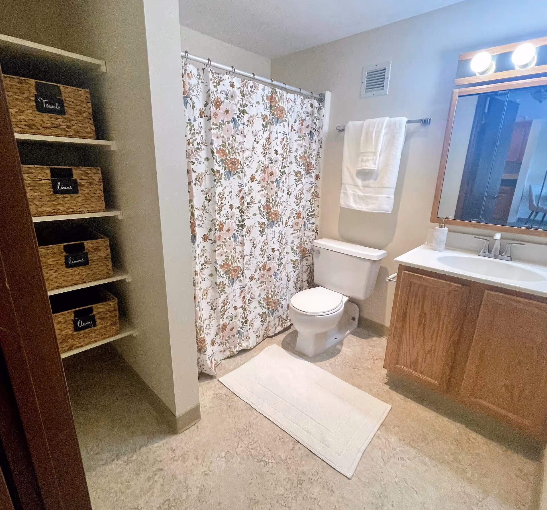 A clean bathroom with a floral shower curtain, a white toilet, a white bath mat on the floor, a wooden vanity with a sink and mirror above it, and shelves with labeled wicker baskets for towels, linens, and cleaning supplies.