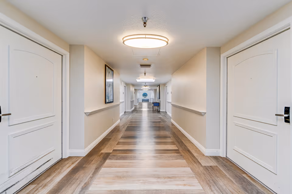 Long, well-lit interior hallway with white doors on both sides and wood-patterned flooring.