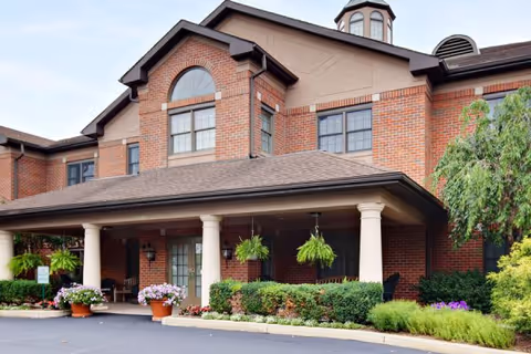 Exterior view of a two-story brick building with a covered entrance supported by white columns. The entrance area is decorated with hanging plants and potted flowers, surrounded by well-maintained shrubs and greenery.