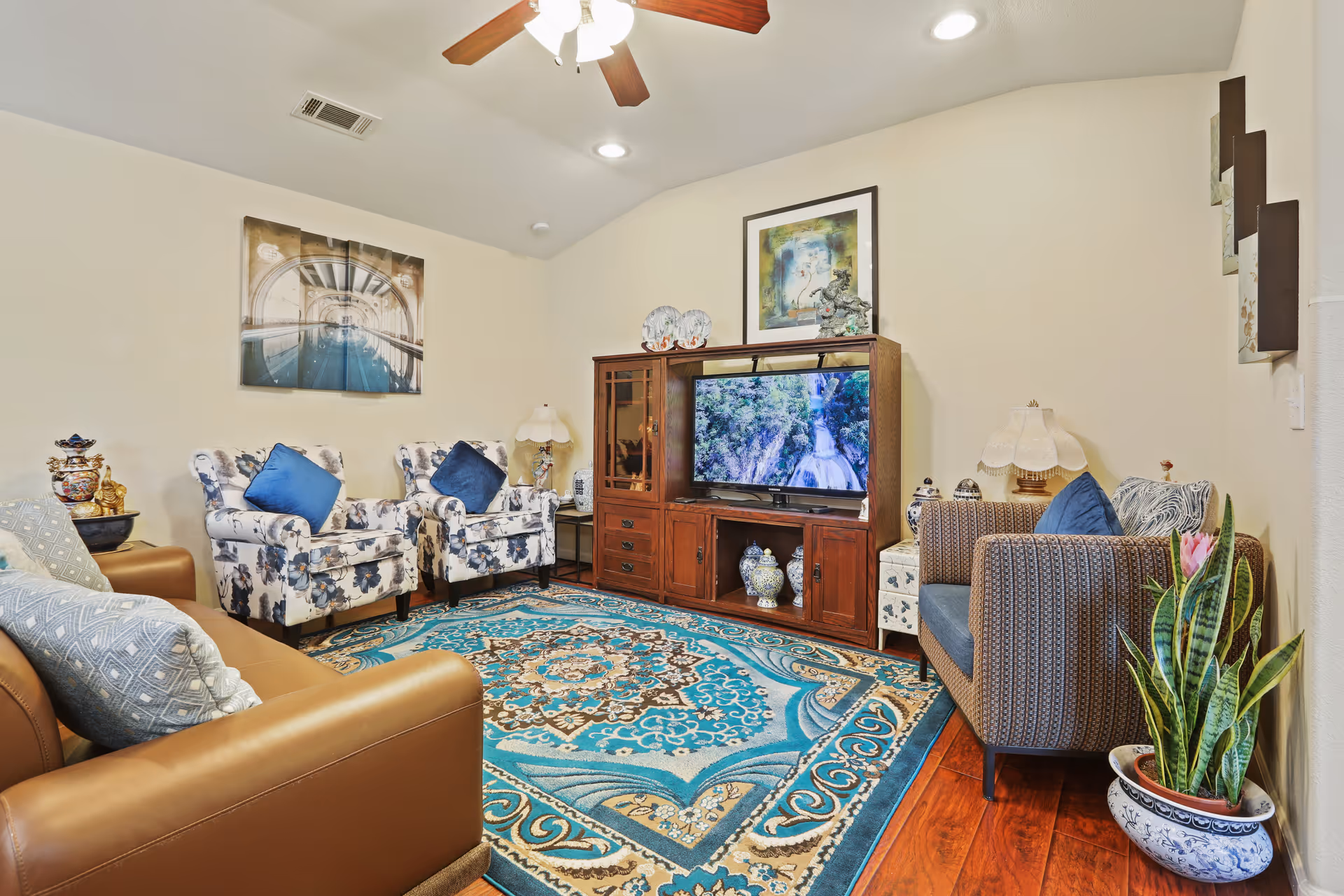 Cozy living room with floral armchairs, a wooden TV cabinet, blue patterned area rug, and a potted plant.