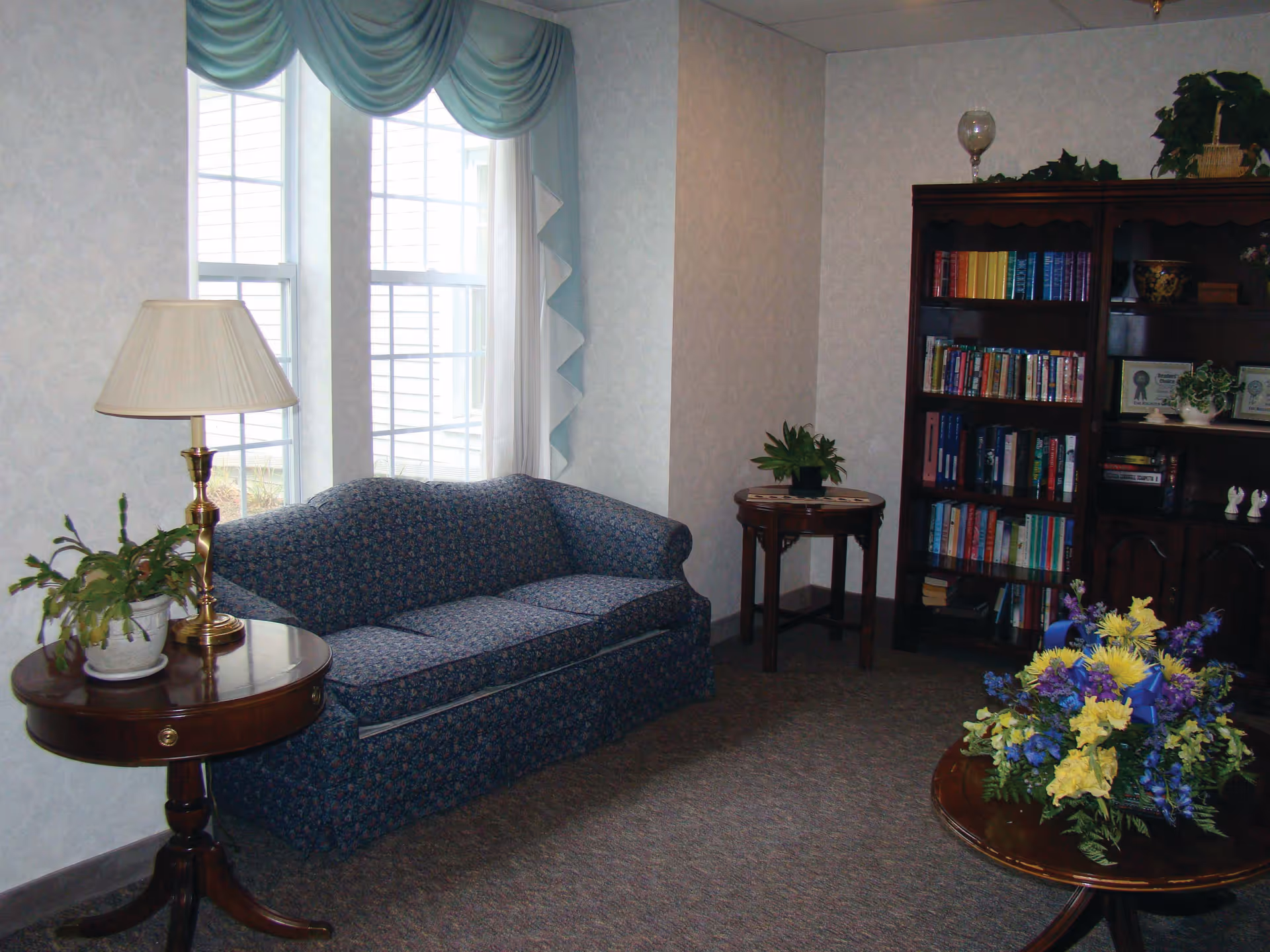 A cozy living room area with a blue patterned sofa, two wooden side tables with potted plants and a lamp, a wooden coffee table with a colorful floral arrangement, and a dark wooden bookshelf filled with books and decorative items. The room has two large windows with light blue valances and white curtains.