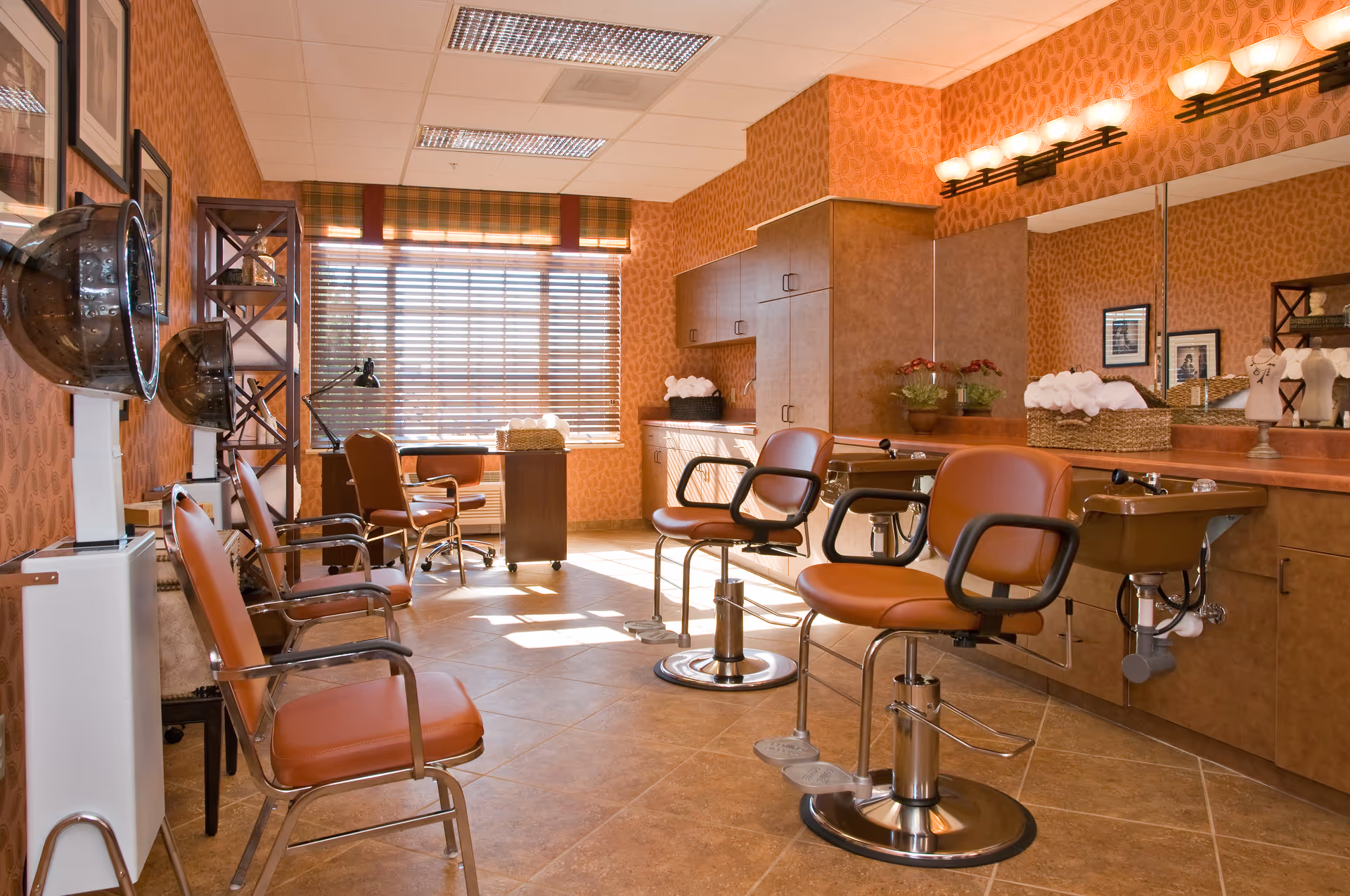 Interior view of a salon area in a senior living facility with multiple brown leather styling chairs, hair dryers, a large mirror, and cabinets. The room has warm orange patterned wallpaper, tiled floor, and a window with blinds letting in natural light.