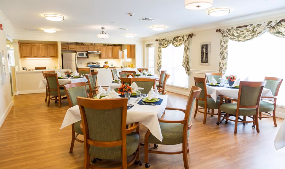 A bright and spacious dining room with several round tables covered with white tablecloths, each set with plates, napkins, glasses, and floral centerpieces. The room has wooden chairs with green upholstery, large windows with floral curtains allowing natural light, and a kitchen area with wooden cabinets and stainless steel appliances in the background.