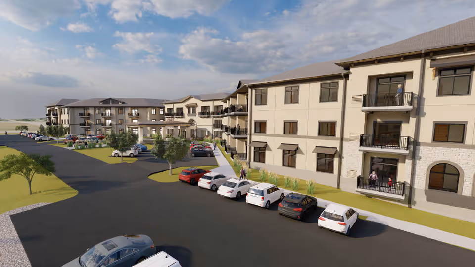 Exterior view of a multi-story senior living facility named The Parkwood with parked cars along the driveway, balconies with people, and landscaped greenery under a partly cloudy sky.