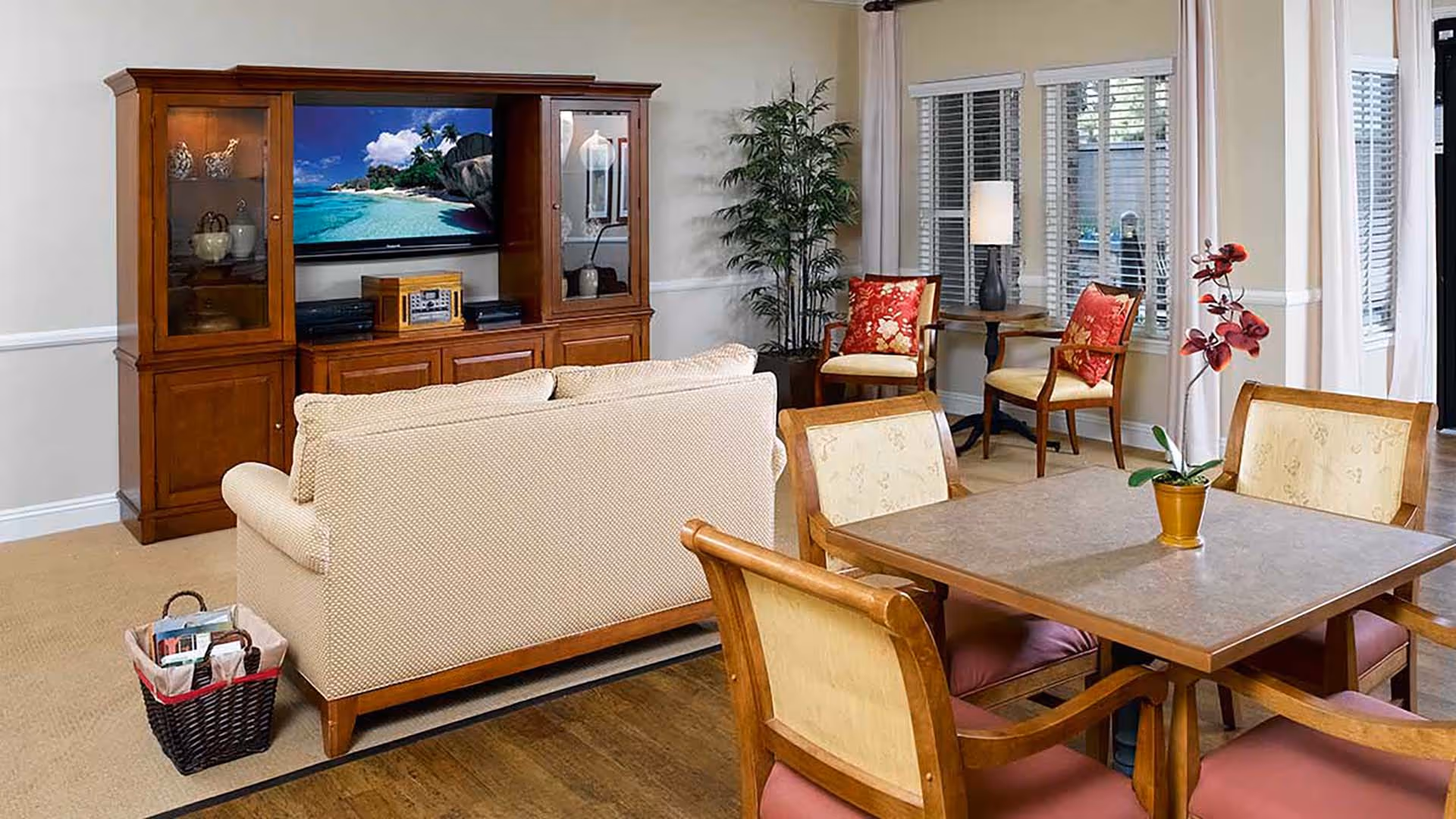 A cozy living room area with a beige sofa facing a wooden entertainment center with a TV displaying a beach scene. There is a basket with magazines next to the sofa. In the foreground, there is a square dining table with four wooden chairs, and a small potted plant with red flowers on the table. In the background, two armchairs with red floral cushions are placed near a window with white blinds and a floor lamp.