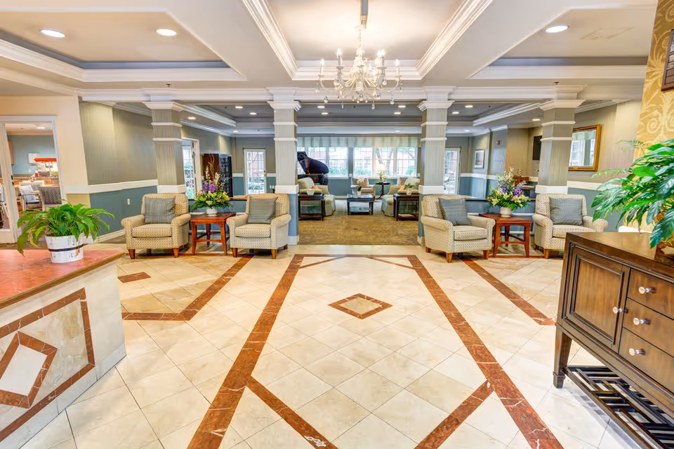 A spacious and well-lit senior living facility common area with tiled floors featuring a decorative pattern, comfortable armchairs arranged around small tables with flower arrangements, large windows letting in natural light, and a chandelier hanging from the ceiling. There is a piano visible in the background and plants placed on counters and tables.