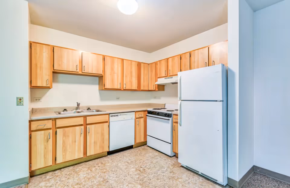 A compact kitchen with light wood cabinets, a sink, a white refrigerator, stove, and dishwasher.